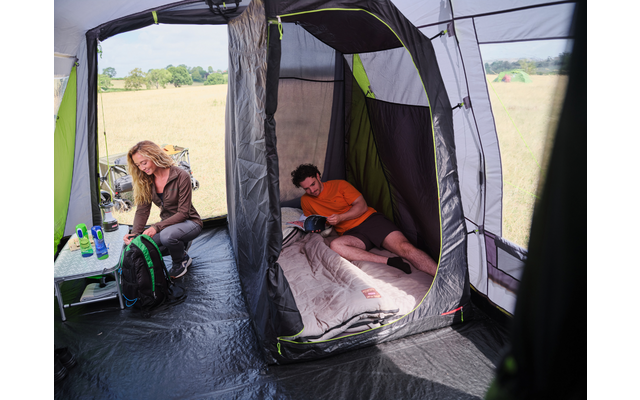 Innenansicht eines Zelts: eine Person liegt in einem Schlafabteil auf einem Schlafsack und hält ein Smartphone; eine weitere Person sitzt auf einem Klappstuhl an einem kleinen Tisch mit Flaschen und einem Rucksack; Feld draußen sichtbar