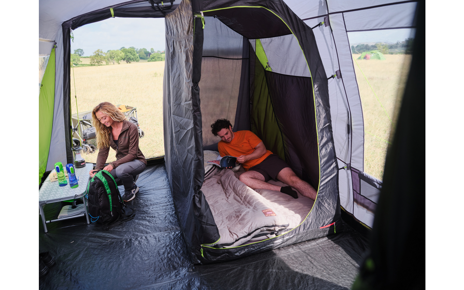 Innenansicht eines Zelts: eine Person liegt in einem Schlafabteil auf einem Schlafsack und hält ein Smartphone; eine weitere Person sitzt auf einem Klappstuhl an einem kleinen Tisch mit Flaschen und einem Rucksack; Feld draußen sichtbar