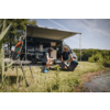 Autocaravana con toldo desplegado en la orilla de un lago. Dos personas arrodilladas en la hierba delante del vehículo junto a una caja abierta y equipo de camping; dos tablas largas en el techo, sillas y una mesa pequeña al lado; orilla y árboles al fondo