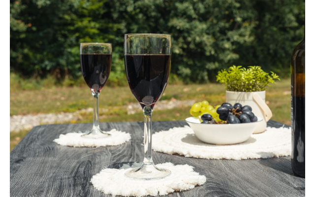 Dos copas de vino con líquido oscuro sobre una mesa de madera oscura, cada una en posavasos redondos blancos; a la derecha una botella de vino, un cuenco con uvas verdes y oscuras y una pequeña maceta con planta; fondo verde desenfocado