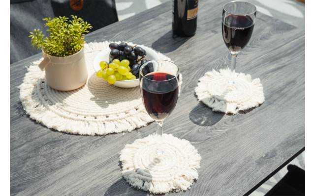Mesa de madera oscura con dos copas de vino tinto sobre posavasos blancos redondos de macramé; junto a ellas un salvamantel redondo de macramé con una maceta pequeña con planta verde, un cuenco de uvas verdes y oscuras y una botella de vino