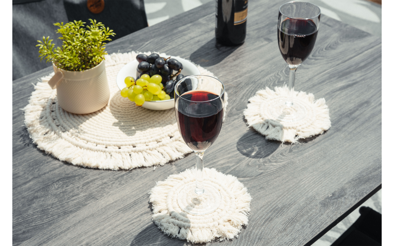 Mesa de madera oscura con dos copas de vino tinto sobre posavasos blancos redondos de macramé; junto a ellas un salvamantel redondo de macramé con una maceta pequeña con planta verde, un cuenco de uvas verdes y oscuras y una botella de vino