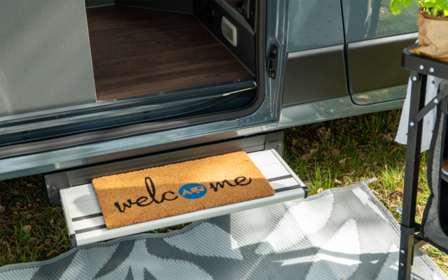 Porte de camping-car ouverte, marche métallique sortie avec paillasson portant l'inscription "welcome", tapis d'extérieur à motifs, herbe et petite table avec plante
