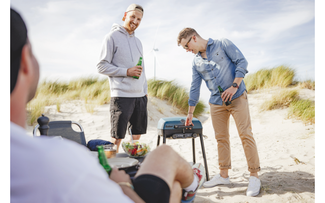 Drei Männer am Strand beim Grillen, einer sitzt, zwei stehen, einer hält ein Bier in der Hand
