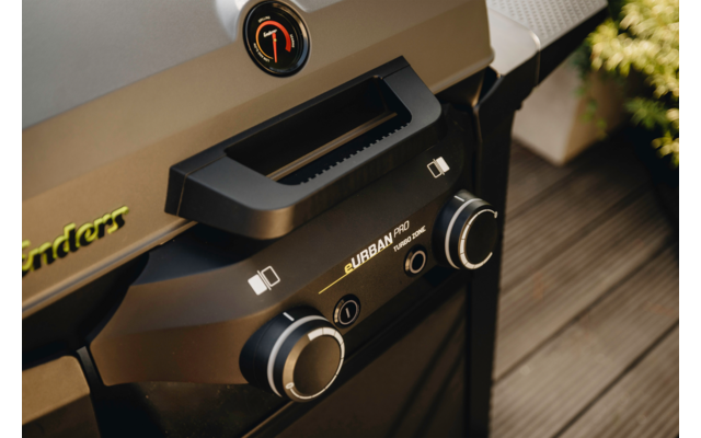 Nahaufnahme eines schwarzen Grills mit Griff, zwei Drehknöpfen, Temperaturanzeige auf dem Deckel, sichtbaren Schriftzügen "Enders" und "URBAN" und einer Pflanze auf einer Holzterrasse