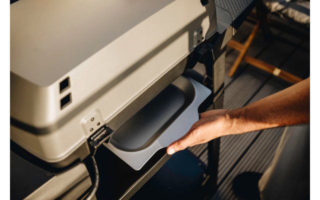 Hand zieht eine flache Schale aus einem hellgrauen Gerät mit zwei rechteckigen Öffnungen; Holzdielenboden und Stuhl im Hintergrund