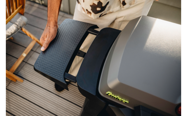 Linke Hand hält einen schwarzen, strukturierten Seitentisch neben einer grauen Grillhaube auf einer Holzterrasse; teilweise sichtbarer Oberkörper und Stuhl im Hintergrund