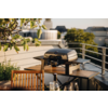Geschlossener Grill auf einem Holztisch auf einer Dachterrasse, mit Holzstuhl, Geländer, Pflanzen und Gebäuden im Hintergrund