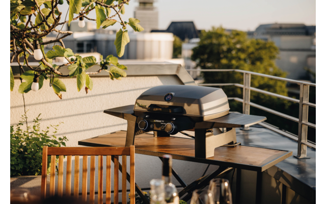 Geschlossener Grill auf einem Holztisch auf einer Dachterrasse, mit Holzstuhl, Geländer, Pflanzen und Gebäuden im Hintergrund