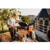 Dachterrasse mit zwei Personen: ein Mann steht an einem geöffneten Grill und hält eine Grillzange, eine Frau sitzt an einem Holztisch mit Gläsern und Tellern; im Hintergrund Stadtgebäude und ein Kirchturm