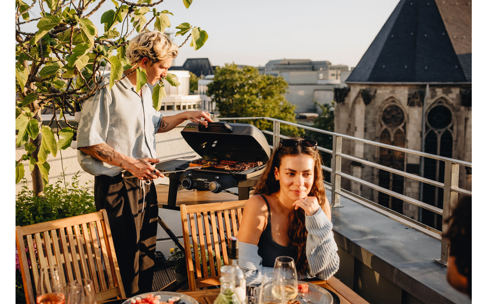 Dachterrasse mit zwei Personen: ein Mann steht an einem geöffneten Grill und hält eine Grillzange, eine Frau sitzt an einem Holztisch mit Gläsern und Tellern; im Hintergrund Stadtgebäude und ein Kirchturm