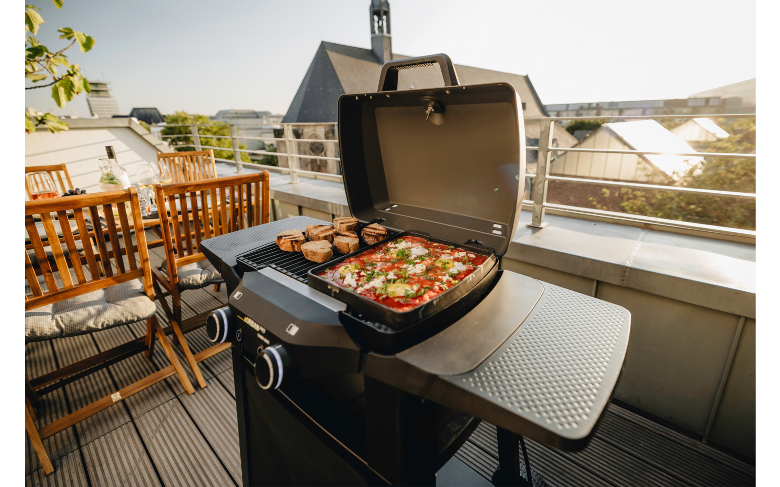 Grill mit offenem Deckel auf einem Balkon; Grillrost mit Gemüse und Brotstücken; Holz-Gartentisch und Stühle neben dem Grill; Dächer und Geländer im Hintergrund