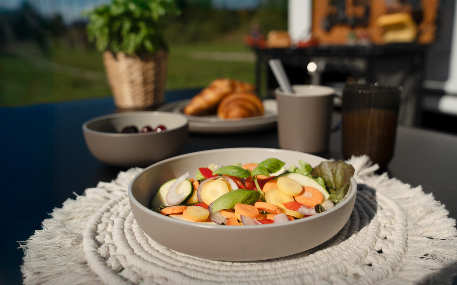 Schüssel mit gemischtem Salat (Gurkenscheiben, Radieschenscheiben, Karottenstreifen, Blattsalat, Basilikumblatt) auf rundem gehäkeltem Platzset; im Hintergrund Croissants auf einem Teller, Schale mit dunklen Früchten, Tasse und Glas; unscharfer Garte