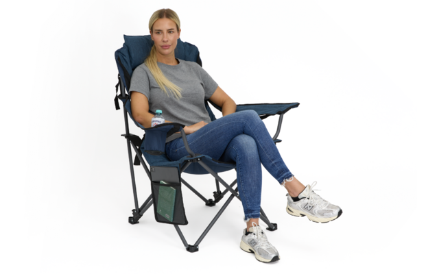 Person sitzt in blauem Klapp-Campingstuhl mit Armlehnen, Seitentasche und Wasserflasche, trägt graues T‑Shirt, blaue Jeans und weiße Sportschuhe, Beine gekreuzt, weißer Hintergrund
