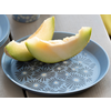 Deux tranches de melon sur une assiette bleue à motif, deux gobelets bleu et blanc en arrière-plan sur une table en bois clair, coussin jaune sur le bord droit