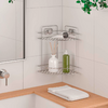 Metal corner shelf mounted on white tiled wall; top shelf with a roll of paper, below a white bottle with reed sticks and a green bottle; left mirror and faucet, wooden counter with folded white towel below, green leaves at top right