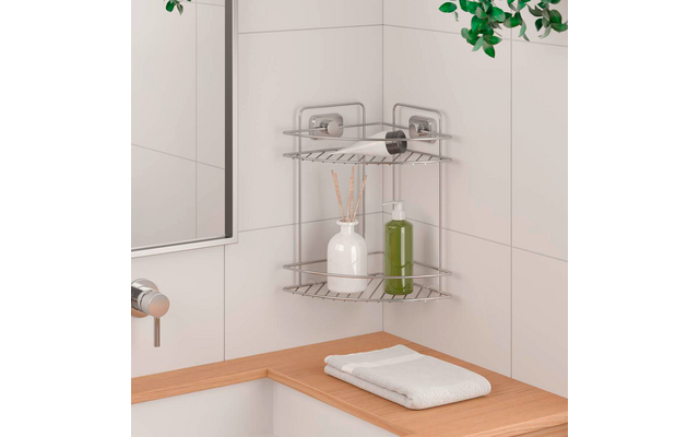 Metal corner shelf mounted on white tiled wall; top shelf with a roll of paper, below a white bottle with reed sticks and a green bottle; left mirror and faucet, wooden counter with folded white towel below, green leaves at top right