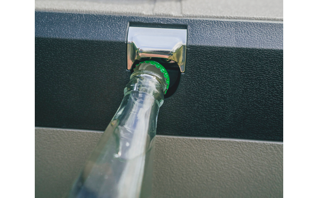 Nahaufnahme einer Glasflasche im Metall-Flaschenöffner, befestigt an einer schwarzen, strukturierten Fläche; grüner Kronkorken sichtbar