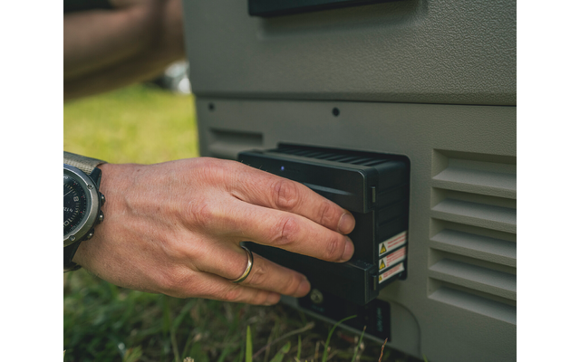 Nahaufnahme einer Hand mit Armbanduhr und Fingerring, die ein schwarzes rechteckiges Modul an ein graues Gerät auf Gras hält