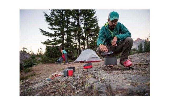 Person in grüner Jacke und Kappe kniet und rührt in einem Topf auf einem kleinen Campingkocher; Zelt und zweite Person sind in der Nähe sichtbar auf felsigem Boden, Nadelbäume und Berge im Hintergrund