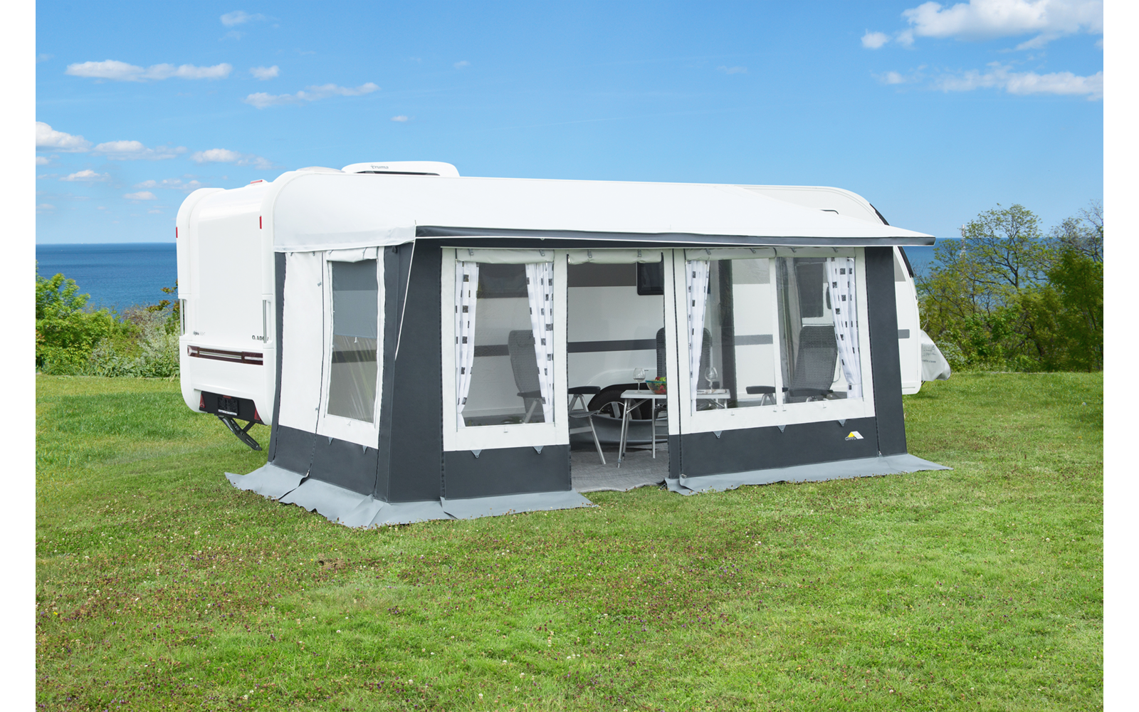 Weißer Wohnwagen mit grauem Vorzelt auf einer grünen Wiese, Meer im Hintergrund und blauer Himmel mit wenigen Wolken; keine Personen sichtbar