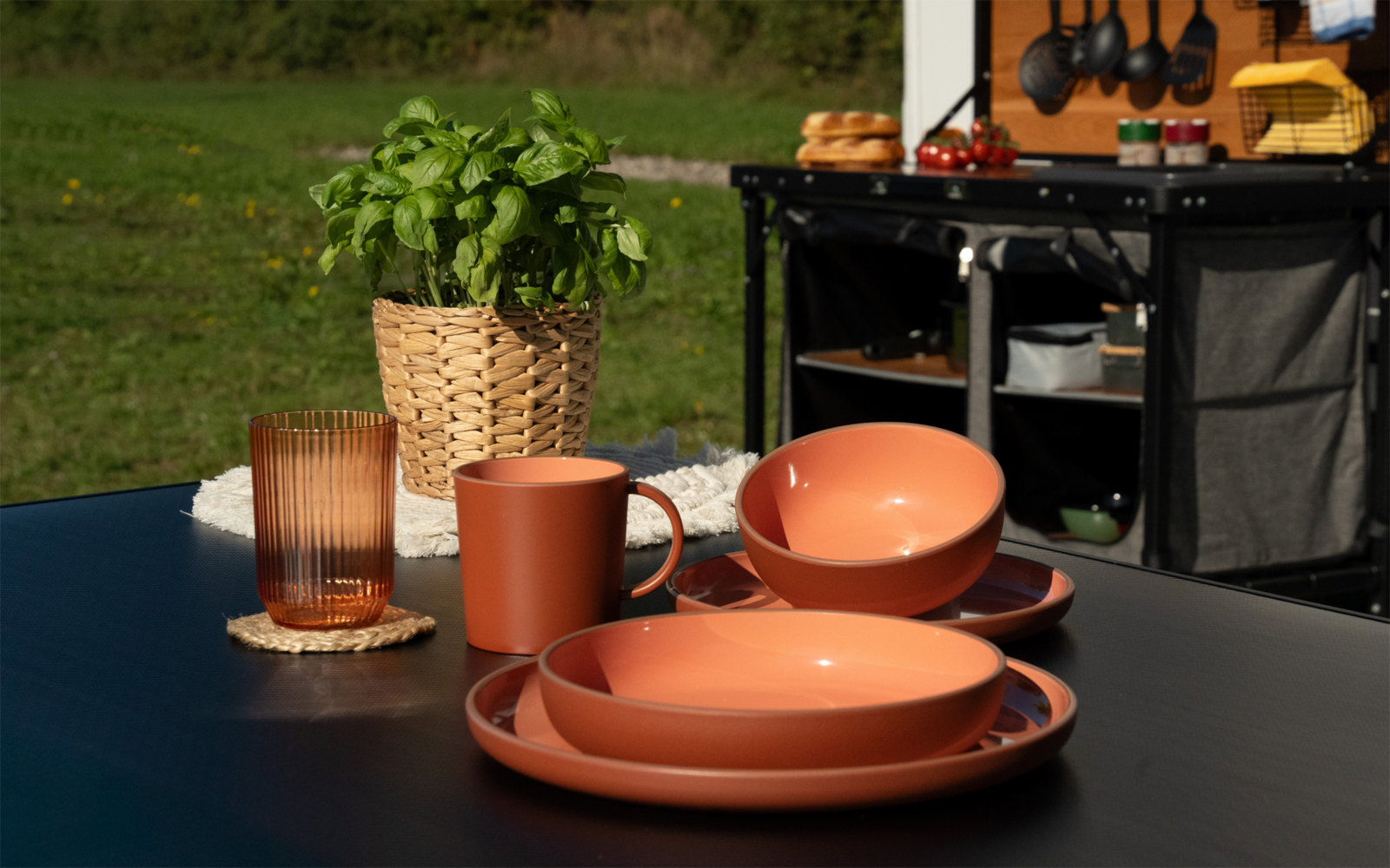 Table de jardin noire avec assiettes, bols et tasse couleur terre cuite empilés, verre cannelé sur dessous-de-verre, plante de basilic dans un panier tressé ; en arrière-plan un chariot de cuisine mobile avec ustensiles et petits pains