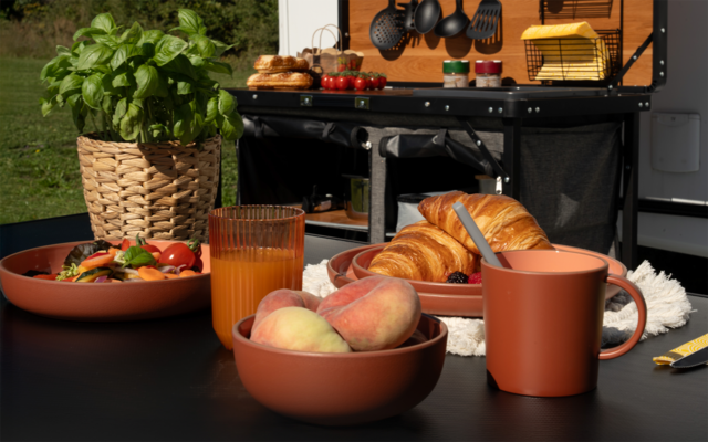Table avec un bol de croissants, un bol de pêches, une tasse, un verre de jus d'orange, un bol de salade, une plante de basilic dans un panier et un chariot de cuisine noir en arrière-plan