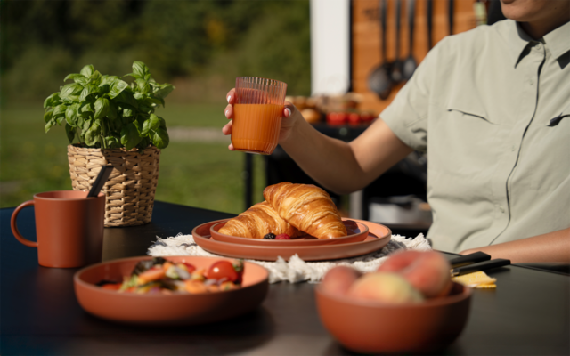 Vue partielle d'une personne en chemise claire tenant un verre contenant un liquide orange; sur une table sombre vaisselle en terre cuite avec deux croissants, un bol de salade avec tomates cerises, un bol de pêches, une tasse, un couteau et un basilic en pot tressé; fond de jardin flou
