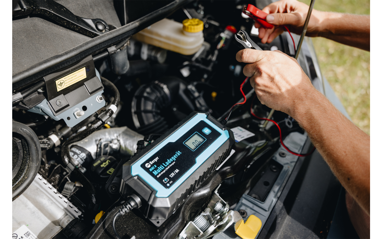 Handen houden rode en zwarte kabelklemmen vast, aangesloten op een zwart-blauw elektronisch apparaat met display in de motorruimte van een auto; motoronderdelen en een gele koelvloeistofdop zichtbaar