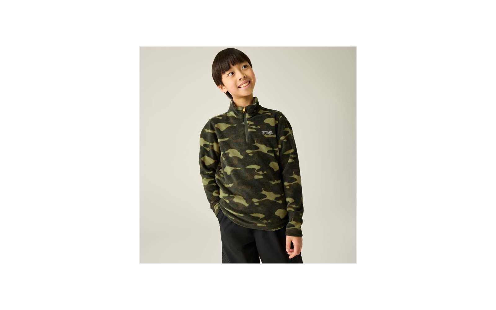 Junge in grünem Camouflage-Fleecepullover mit halbem Reißverschluss, Hände in den Taschen, schaut nach oben rechts vor hellem Hintergrund