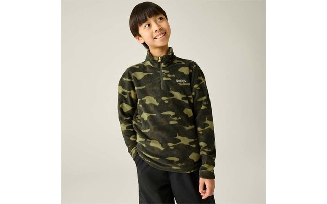 Junge in grünem Camouflage-Fleecepullover mit halbem Reißverschluss, Hände in den Taschen, schaut nach oben rechts vor hellem Hintergrund