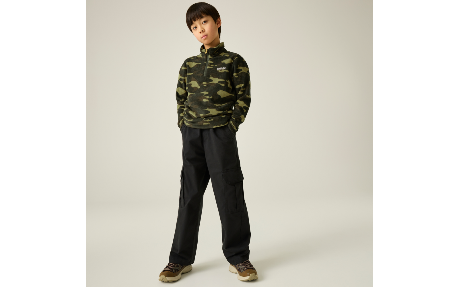 Ein Kind vor hellem einfarbigem Hintergrund, trägt grünen Camouflage‑Pullover mit Kapuze, schwarze Cargohose und braune Sneakers, Hände in den Taschen