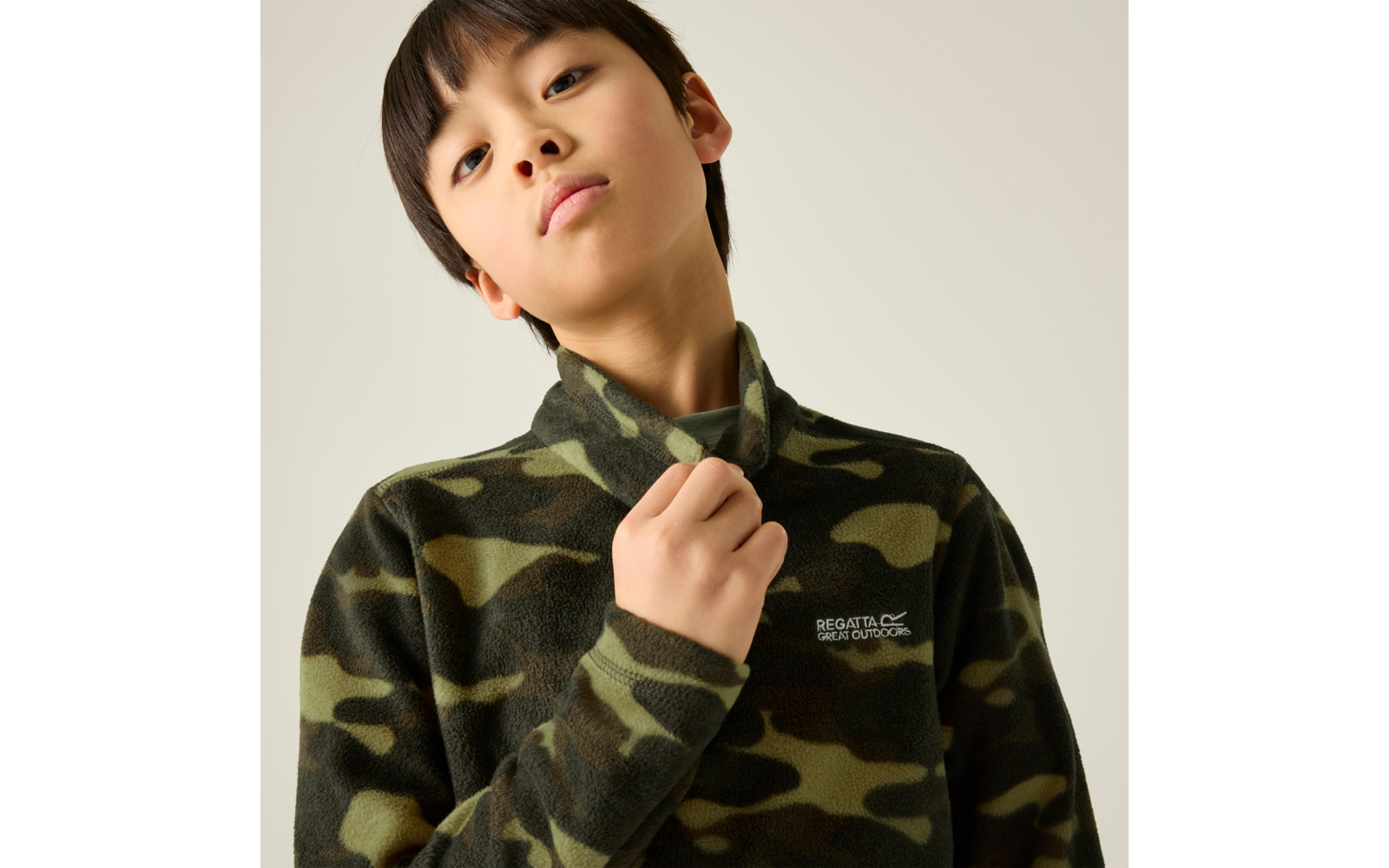 Kind mit kurzem dunklem Haar trägt olivgrünes Camouflage-Fleece, hält den Kragen, heller Hintergrund