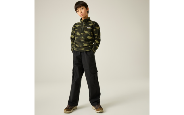 Ein Kind vor hellem einfarbigem Hintergrund, trägt grünen Camouflage‑Pullover mit Kapuze, schwarze Cargohose und braune Sneakers, Hände in den Taschen