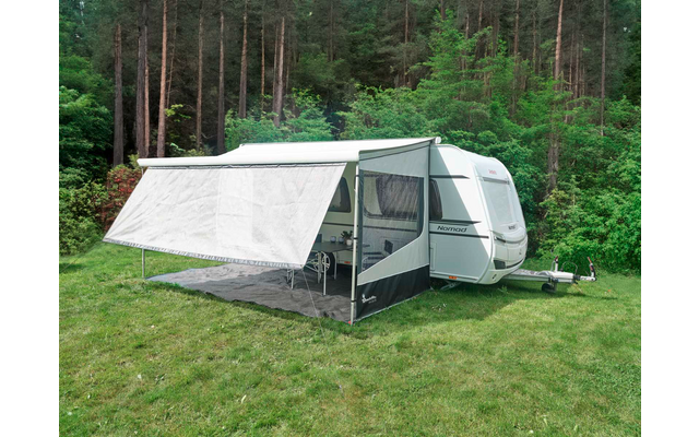 Caravana blanca con toldo blanco instalado sobre césped verde frente a un bosque de pinos; alfombra de suelo y sillas de camping visibles