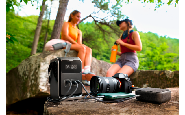 Draagbare powerbank, camera met lens, smartphone en oplaadkabels op een rots; op de achtergrond twee personen in sportkleding, één houdt een drinkfles vast, bomen en groen