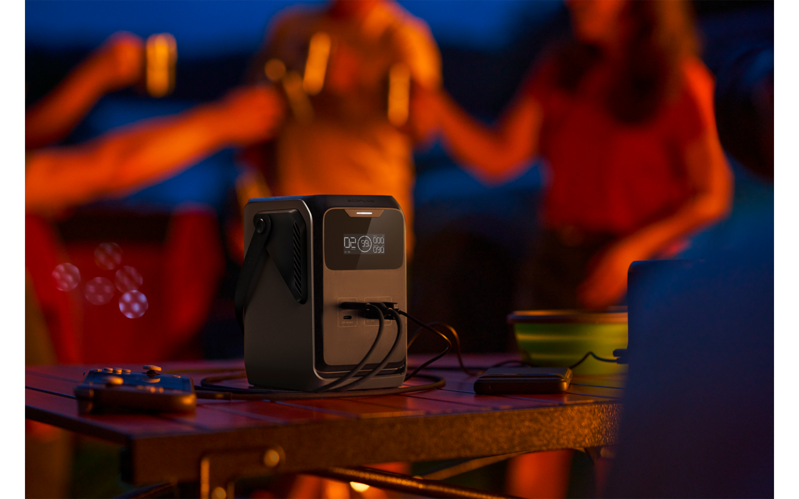 Zwarte draagbare powerstation met display en twee aangesloten kabels op een tafel; op de achtergrond vage personen met mokken