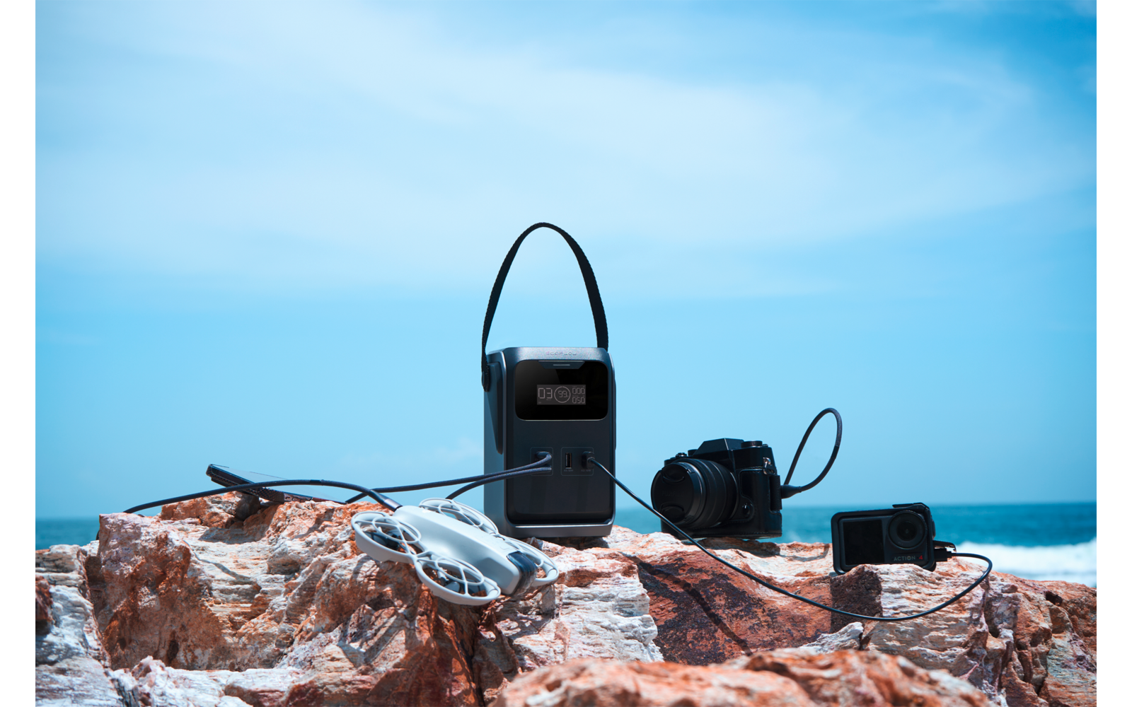 Op een rotsachtig uitsteeksel bij de zee: een zwart apparaat met draaghandgreep, een drone met propellers, een camera en een actioncam verbonden met kabels; zee en blauwe lucht op de achtergrond