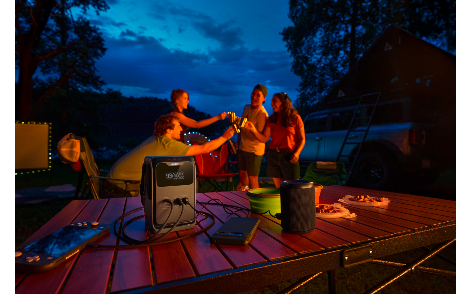 Vijf personen bij een campingtafel in de schemering, ze proosten met drankjes. Op de tafel: een draagbare stroomunit met kabels, een luidspreker en borden met snacks. Op de achtergrond: camper, bomen en donkerblauwe lucht