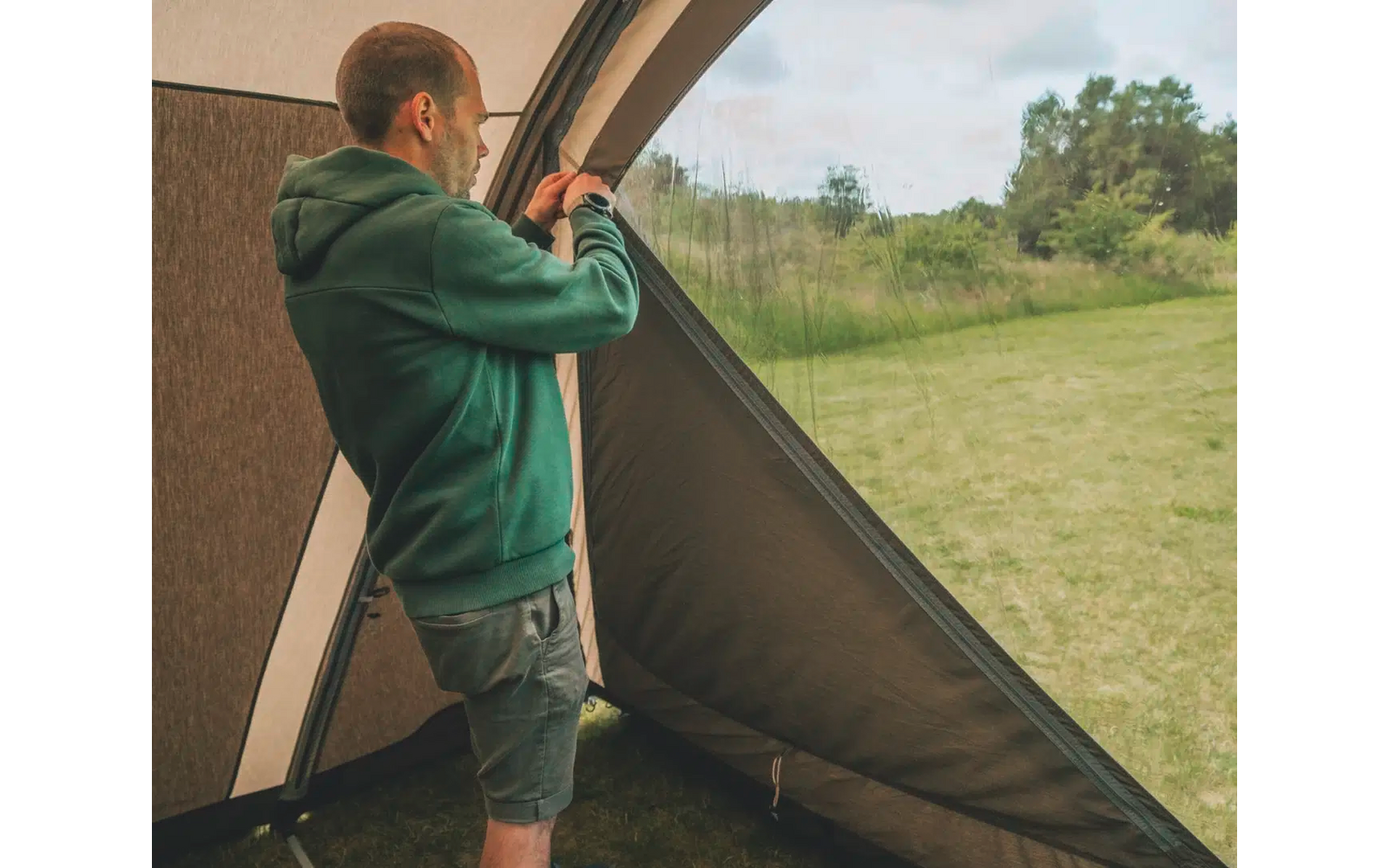 Mann in grünem Pullover schaut durch Teleskop aus einem Zelt auf eine grüne Wiese