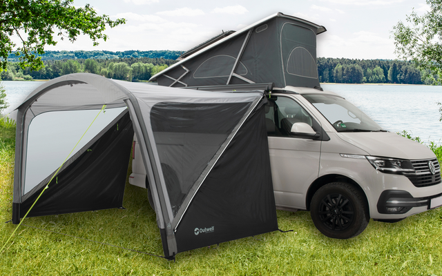 Furgoneta blanca con tienda de techo desplegada y toldo cerrado en un prado verde cerca de un lago con árboles al fondo