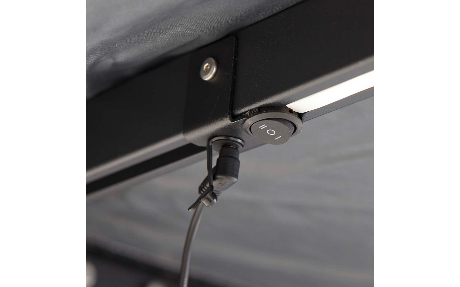 Nahaufnahme eines schwarzen Metallrahmens mit rundem schwarzen Knopf mit den Zeichen 'I' und 'O', angeschlossenem rechtwinkligen schwarzen Stecker mit herabhängendem Kabel, schmaler LED-Leiste und sichtbarer Schraube auf grauem Stoffhintergrund