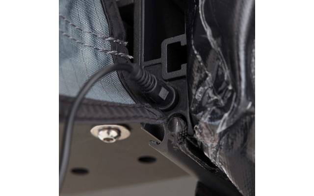 Nahaufnahme eines schwarzen zylindrischen Steckers in einer Buchse an einem schwarzen Kunststoffrahmen, daneben grauer, genähter Stoff und eine silberne Schraube