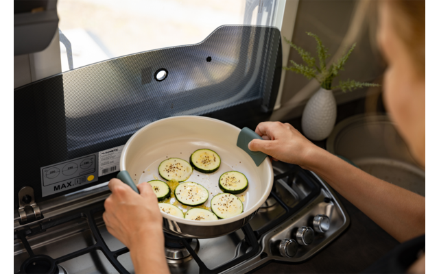 Dos manos sostienen una sartén blanca con asas verdes que contiene rodajas de calabacín con condimentos sobre una cocina de gas; ventana y pequeña planta en maceta al fondo