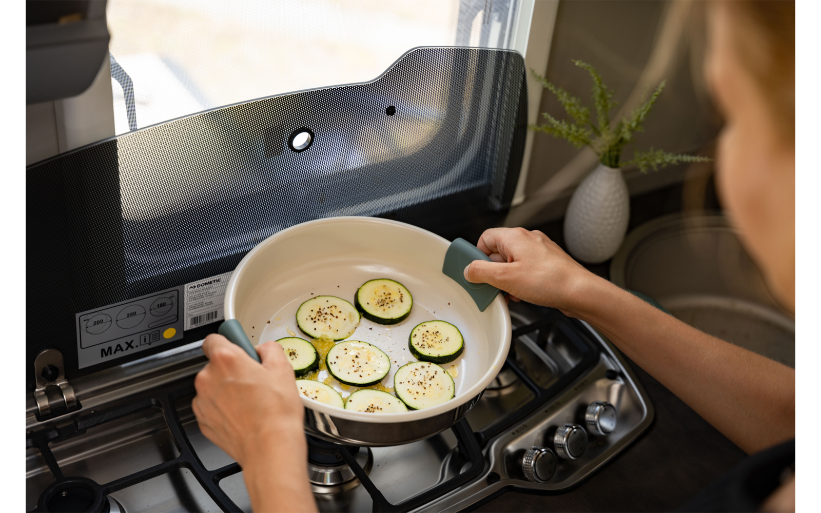 Dos manos sostienen una sartén blanca con asas verdes que contiene rodajas de calabacín con condimentos sobre una cocina de gas; ventana y pequeña planta en maceta al fondo