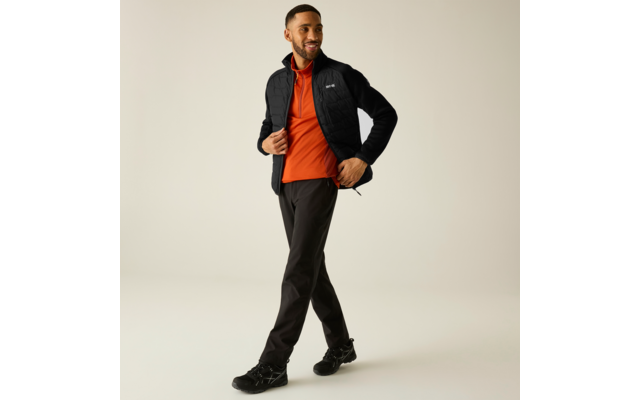 Mann in schwarzer Steppjacke, orangefarbenem Pullover, schwarzen Hosen und schwarzen Sneakers geht vor beigem Hintergrund und lächelt