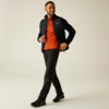 Mann in schwarzer Steppjacke, orangefarbenem Pullover, schwarzen Hosen und schwarzen Sneakers geht vor beigem Hintergrund und lächelt