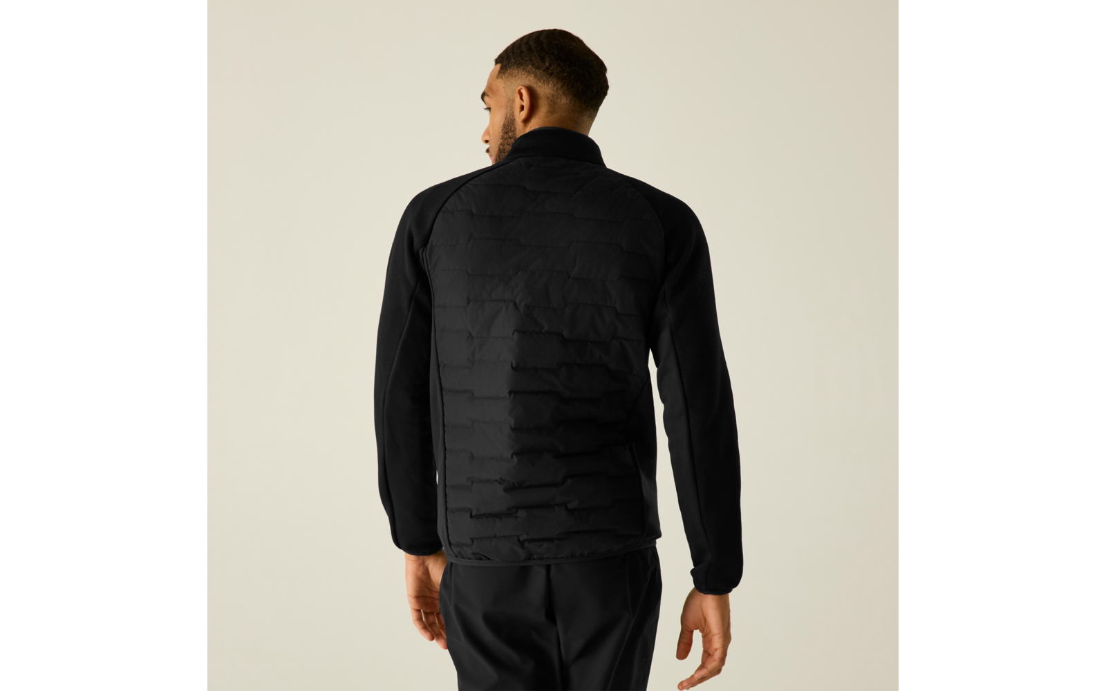 Person von hinten mit kurzem Haar, schwarze gesteppte Jacke und schwarze Hose vor hellem beige Hintergrund
