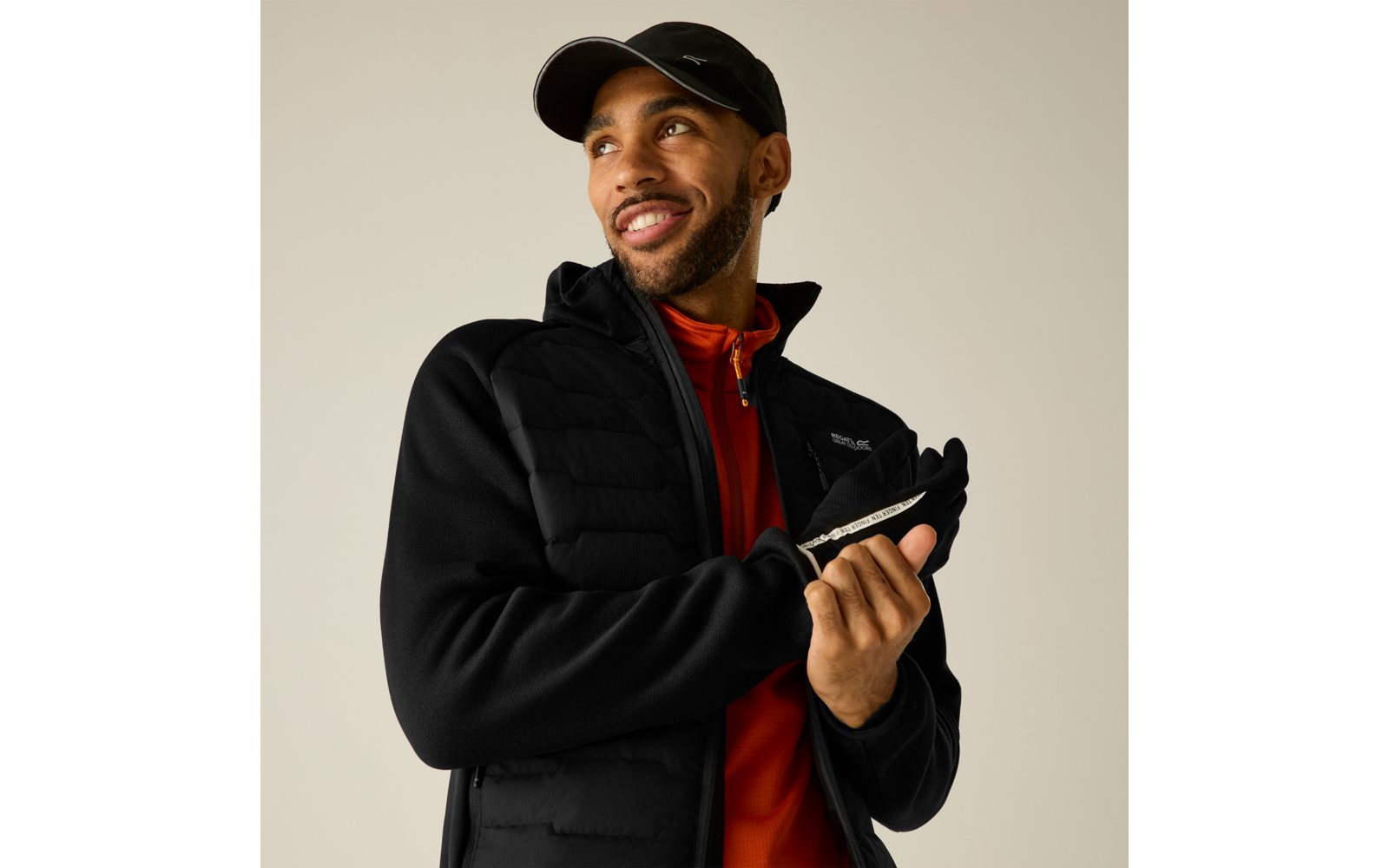 Person mit schwarzer Kappe, schwarzer gesteppter Jacke und oranger Zip-Up zieht einen Handschuh an, Blick nach oben rechts, neutraler Hintergrund