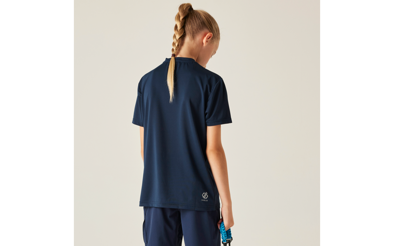 Person von hinten mit geflochtener blonder Zopf, trägt dunkelblaues kurzärmeliges Shirt und dunkelblaue Shorts, hält ein blau-schwarzes Seil in der rechten Hand vor beigem Hintergrund